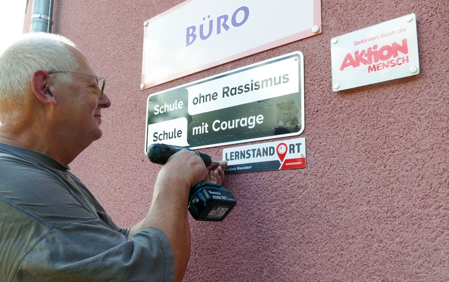 Ein älterer Mann befestigt mit einer Bohrmaschine ein Schild an einer Wand, auf dem die Aufschriften „Schule ohne Rassismus“, „Schule mit Courage“ und „Lernstandort“ zu lesen sind. An der Wand befinden sich außerdem Schilder mit den Aufschriften „Büro“ und „Aktion Mensch“.