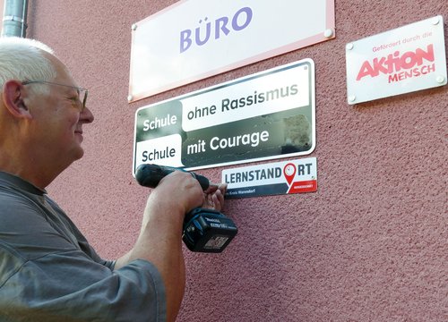 Ein älterer Mann befestigt mit einer Bohrmaschine ein Schild an einer Wand, auf dem die Aufschriften „Schule ohne Rassismus“, „Schule mit Courage“ und „Lernstandort“ zu lesen sind. An der Wand befinden sich außerdem Schilder mit den Aufschriften „Büro“ und „Aktion Mensch“.