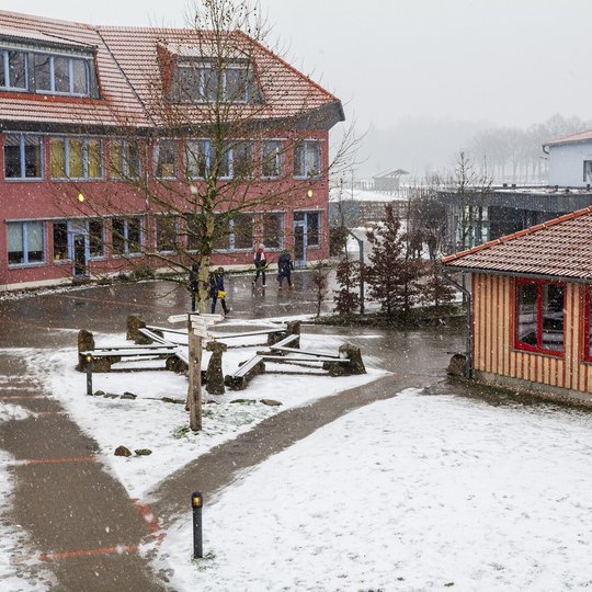 An einem verschneiten Tag gehen Schüler draußen zwischen Schulgebäuden mit roten Dächern hindurch, während Schnee den Boden, die Bäume und die Bänke auf dem Schulhof bedeckt.