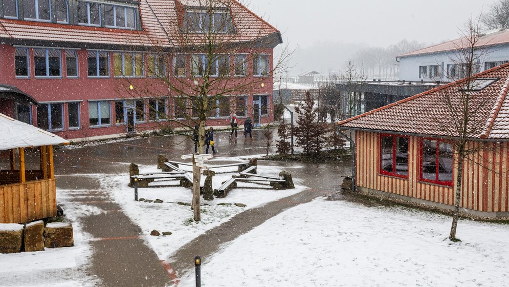 An einem verschneiten Tag gehen Schüler draußen zwischen Schulgebäuden mit roten Dächern hindurch, während Schnee den Boden, die Bäume und die Bänke auf dem Schulhof bedeckt.
