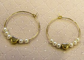 Zwei goldene Creolen mit kleinen weißen Perlen und goldenen herzförmigen Perlen liegen auf einem strukturierten, hellrosa Stoff mit Rautenmuster.