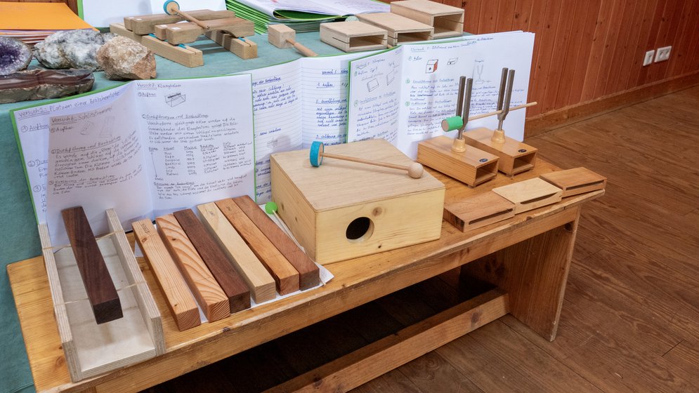 Auf einem Holztisch in einem Klassenzimmer mit Holzwänden und -boden sind Lernhilfen aus Holz, Bausteine und wissenschaftliche Notizen ausgestellt. Im Hintergrund sind verschiedene Instrumente und handschriftliche Notizen zu sehen.