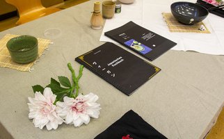 Ein Tisch, auf dem Utensilien für die japanische Teezeremonie angerichtet sind: zwei rosa Blumen, eine grüne Teetasse, eine schwarze Schale und eine Broschüre mit dem Titel „Die japanische Teezeremonie“.