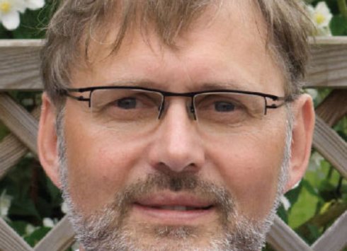 Ein Mann mittleren Alters mit Brille, kurzen braunen Haaren und einem ergrauten Bart steht im Freien vor einem hölzernen Spalier, hinter dem sich grünes Laub und weiße Blüten abzeichnen.