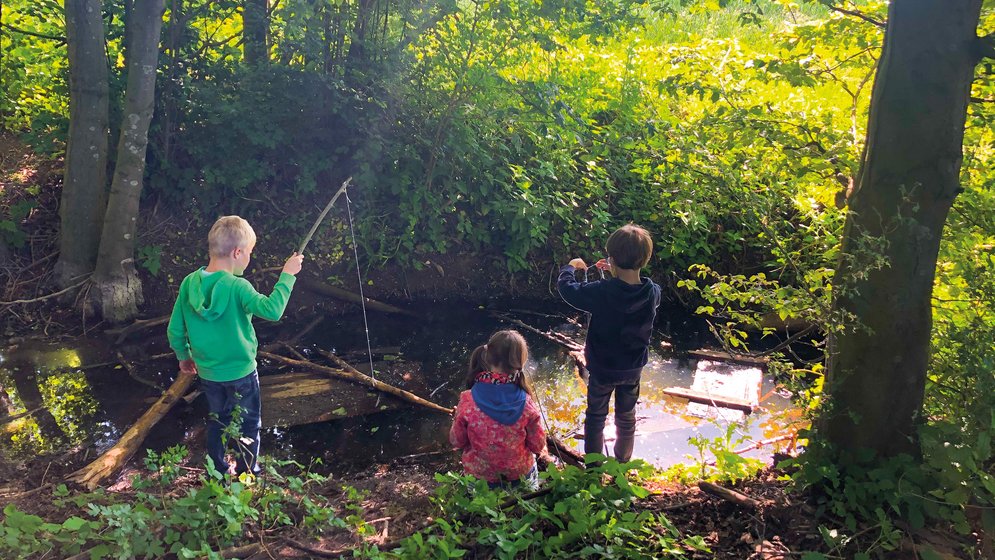 Drei Kinder stehen an einem kleinen, von Bäumen gesäumten Bach. Zwei halten Stöcke über das Wasser, als würden sie angeln, während das dritte am Ufer kniet, umgeben von grünem Laub und Sonnenlicht, das durch die Bäume fällt.