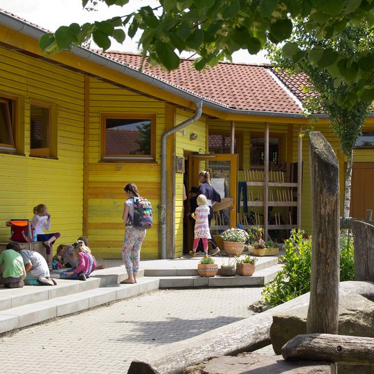 Kinder und Erwachsene versammeln sich vor einem gelben Schulgebäude, vor dem eine Tafel mit Kreidezeichnungen, Topfpflanzen und Holzspielgeräte unter einem blättrigen Baum stehen.