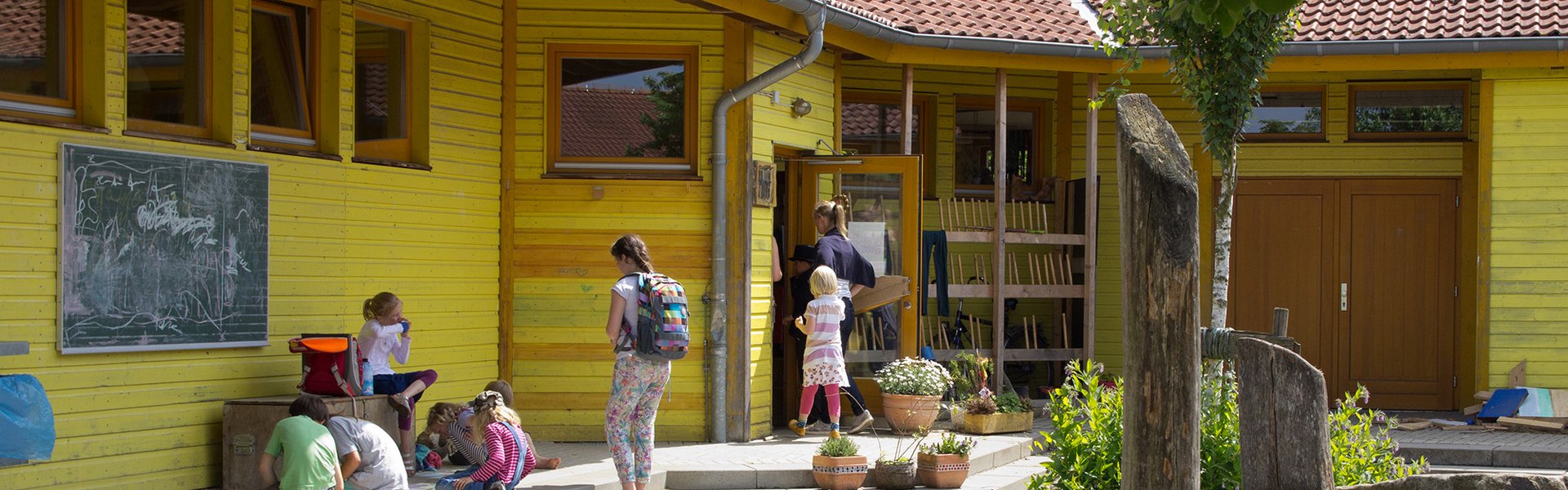 Kinder und Erwachsene versammeln sich vor einem gelben Schulgebäude, vor dem eine Tafel mit Kreidezeichnungen, Topfpflanzen und Holzspielgeräte unter einem blättrigen Baum stehen.