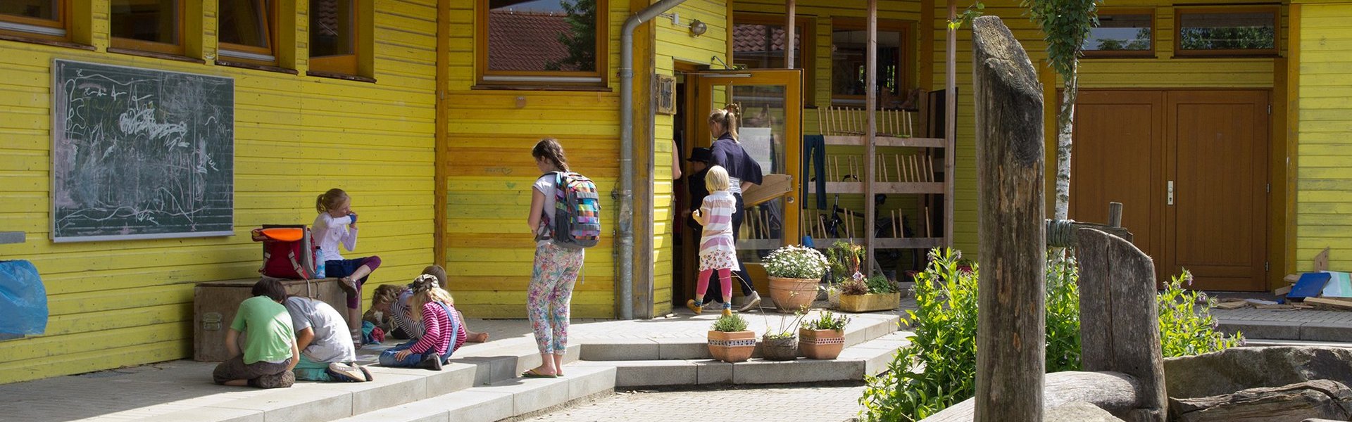 Kinder und Erwachsene versammeln sich vor einem gelben Schulgebäude, vor dem eine Tafel mit Kreidezeichnungen, Topfpflanzen und Holzspielgeräte unter einem blättrigen Baum stehen.