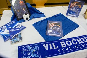 Auf einem Tisch sind Fanartikel des VfL Bochum ausgestellt: ein Schal, ein Mini-Fußball, Vereinsbücher, ein gerahmtes Foto von drei Spielern, ein blaues Tuch und Broschüren, alles ordentlich auf einer weißen Tischdecke angeordnet.