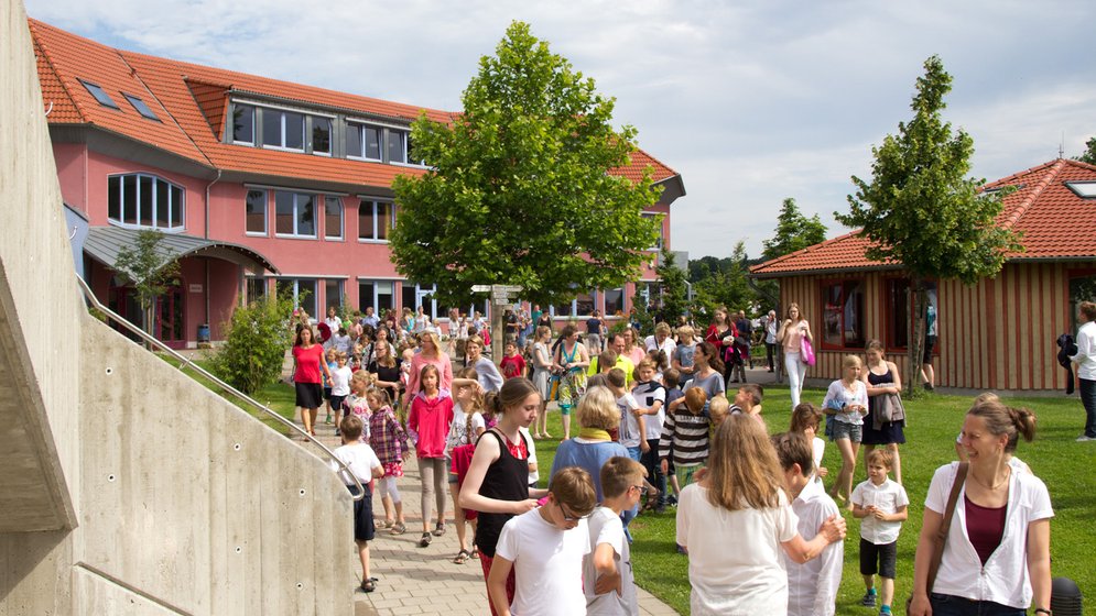 An einem sonnigen Tag versammelt sich eine große Gruppe von Kindern und Erwachsenen vor einem Schulgebäude; sie spazieren und unterhalten sich auf Wegen, die von grünem Gras und Bäumen gesäumt sind.