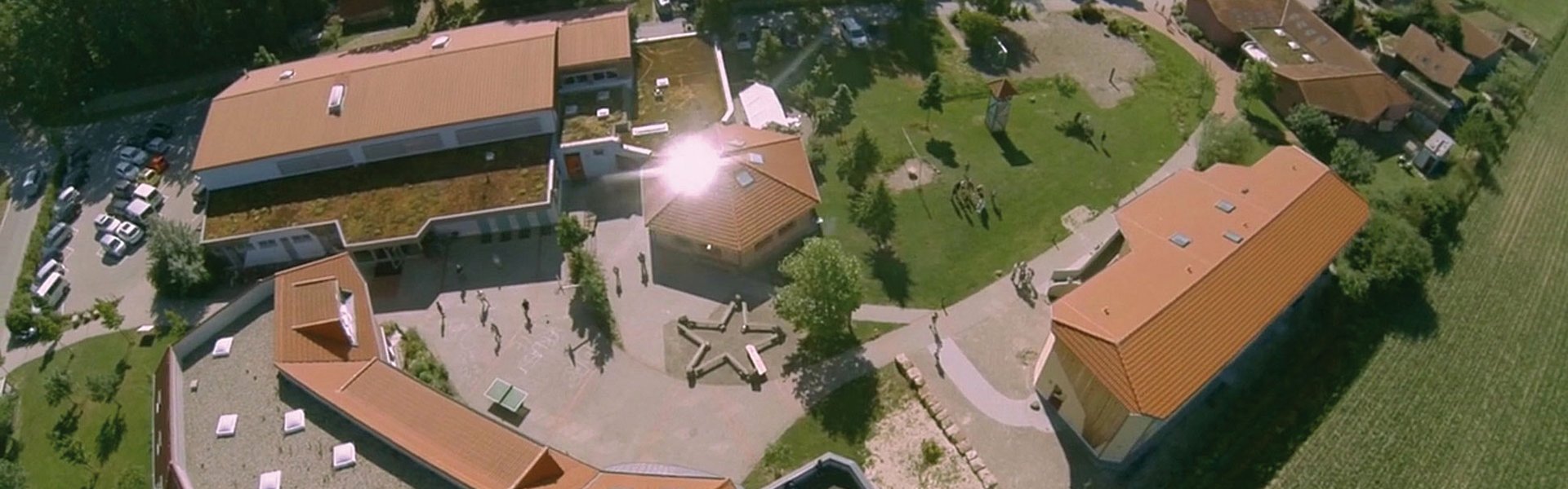 Luftaufnahme eines Schulgeländes mit Gebäuden mit roten Dächern, grünen Rasenflächen, Bäumen und Menschen, die an einem sonnigen Tag draußen spazieren gehen.