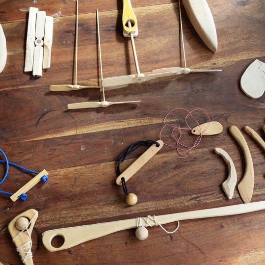 Auf einem Holztisch liegen verschiedene handgefertigte Holzspielzeuge und -gegenstände verstreut, darunter Kreisel, Herzen, Steinschleudern und andere Spielsachen; an einigen sind Schnüre oder Gummibänder befestigt.