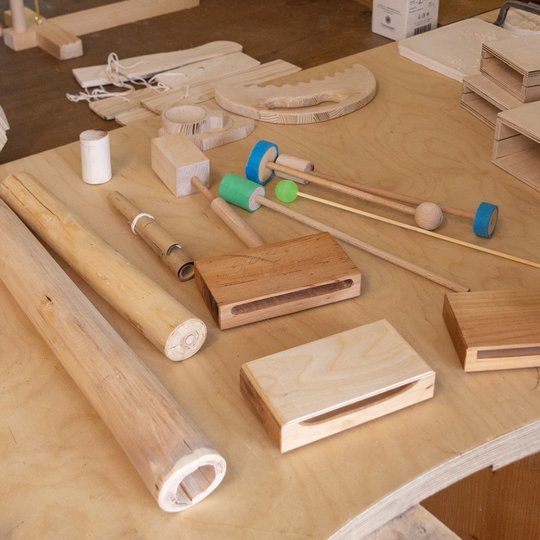 Auf einem hellen Holztisch sind verschiedene Holzgegenstände und -instrumente angeordnet; dabei sind die Hände zweier Personen zu sehen, die mit den Materialien arbeiten.