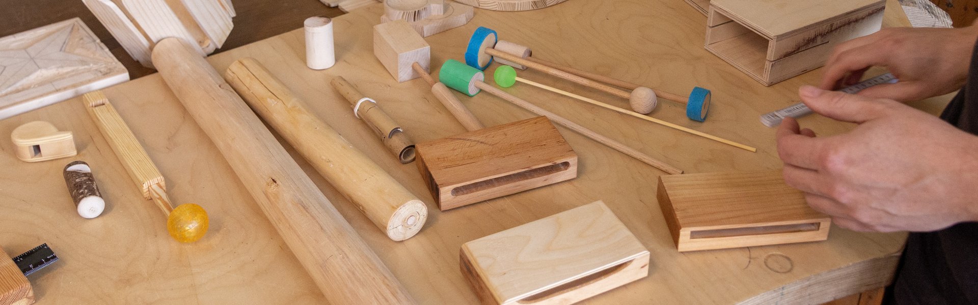 Auf einem hellen Holztisch sind verschiedene Holzgegenstände und -instrumente angeordnet; dabei sind die Hände zweier Personen zu sehen, die mit den Materialien arbeiten.