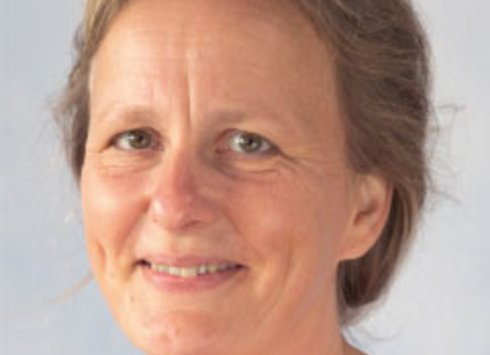 Eine Frau mittleren Alters mit hellbraunen Haaren, die sanft lächelt, ein rotes Oberteil trägt und vor einem schlichten, hellen Hintergrund posiert.