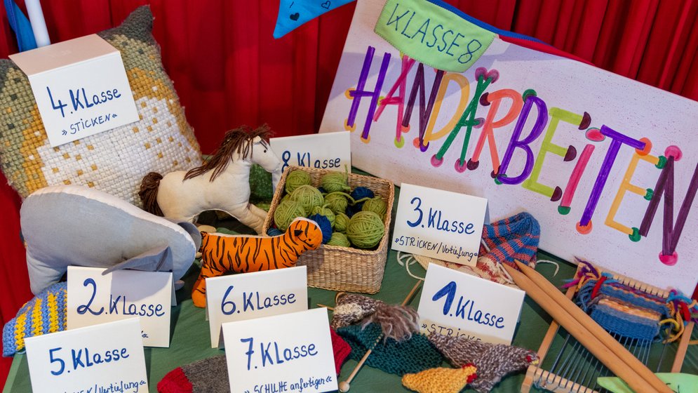 Eine farbenfrohe Ausstellung verschiedener handgefertigter Kunsthandwerksartikel, Garn und Strickwaren mit Schildern, auf denen verschiedene Schulklassen und das Wort „Handarbeiten“ auf Deutsch zu sehen sind, vor einem roten Vorhang als Hintergrund.