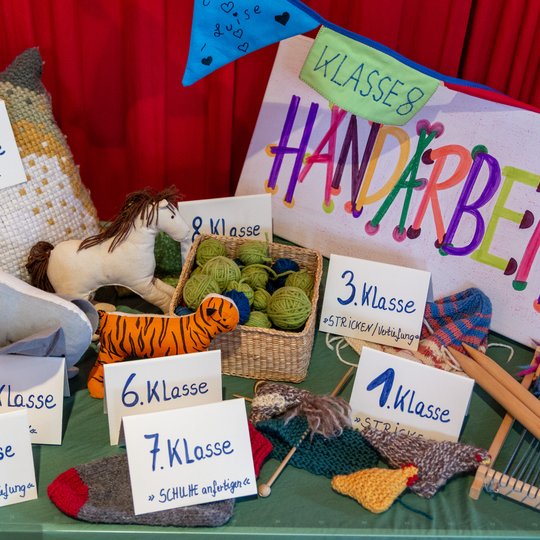 Eine farbenfrohe Ausstellung verschiedener handgefertigter Kunsthandwerksartikel, Garn und Strickwaren mit Schildern, auf denen verschiedene Schulklassen und das Wort „Handarbeiten“ auf Deutsch zu sehen sind, vor einem roten Vorhang als Hintergrund.