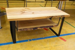Ein Couchtisch aus Holz mit naturbelassener Kante, schwarzen Metallbeinen und einer unteren Ablage, der auf einem gelben Boden mit farbigen Linien steht.