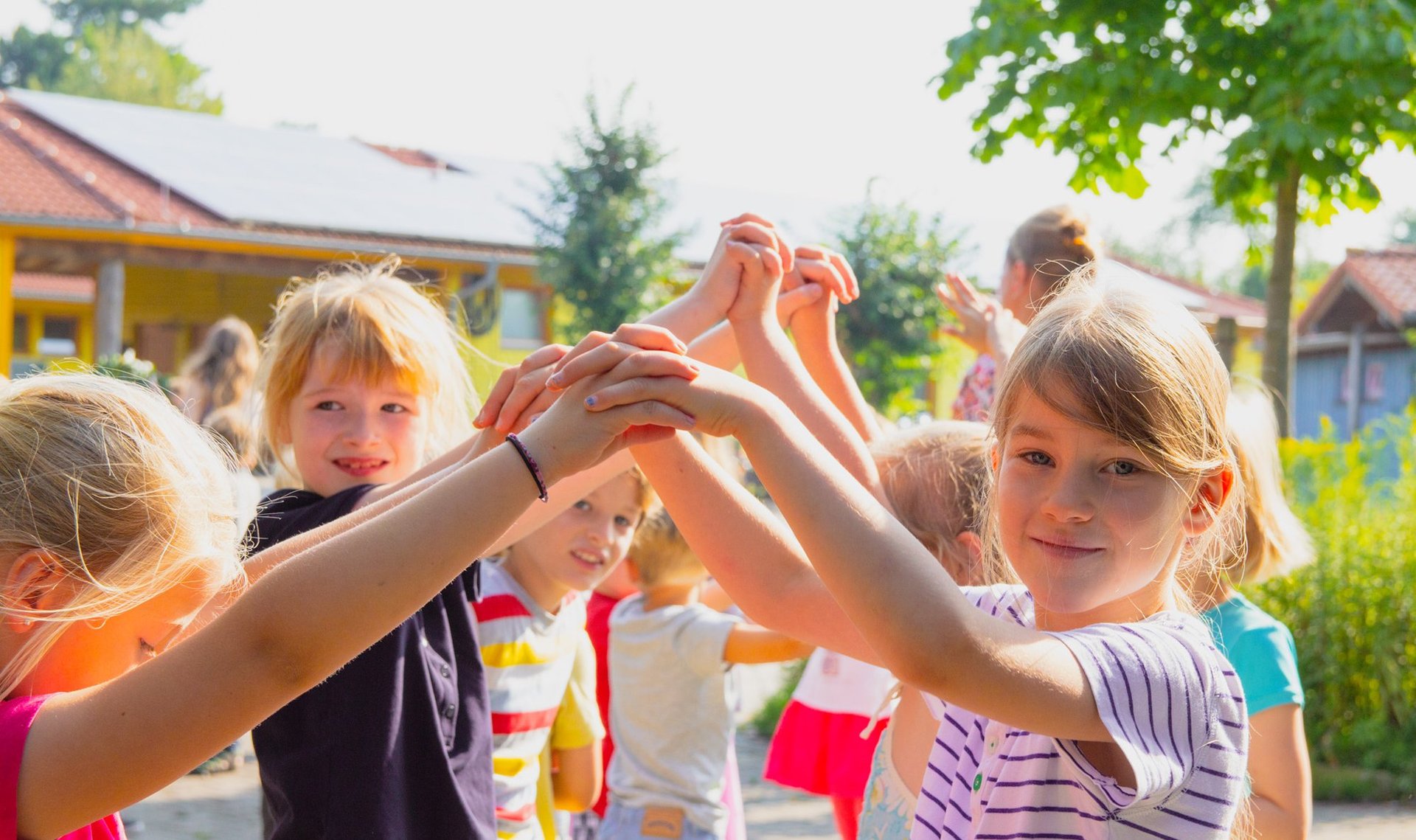 Kinder spielen draußen, lächeln und halten sich an den Händen, während sie im Kreis stehen; im Hintergrund sind Bäume und Gebäude zu sehen – an einem sonnigen Tag.