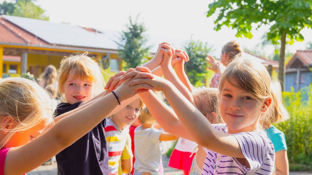 Kinder spielen draußen, lächeln und halten sich an den Händen, während sie im Kreis stehen; im Hintergrund sind Bäume und Gebäude zu sehen – an einem sonnigen Tag.