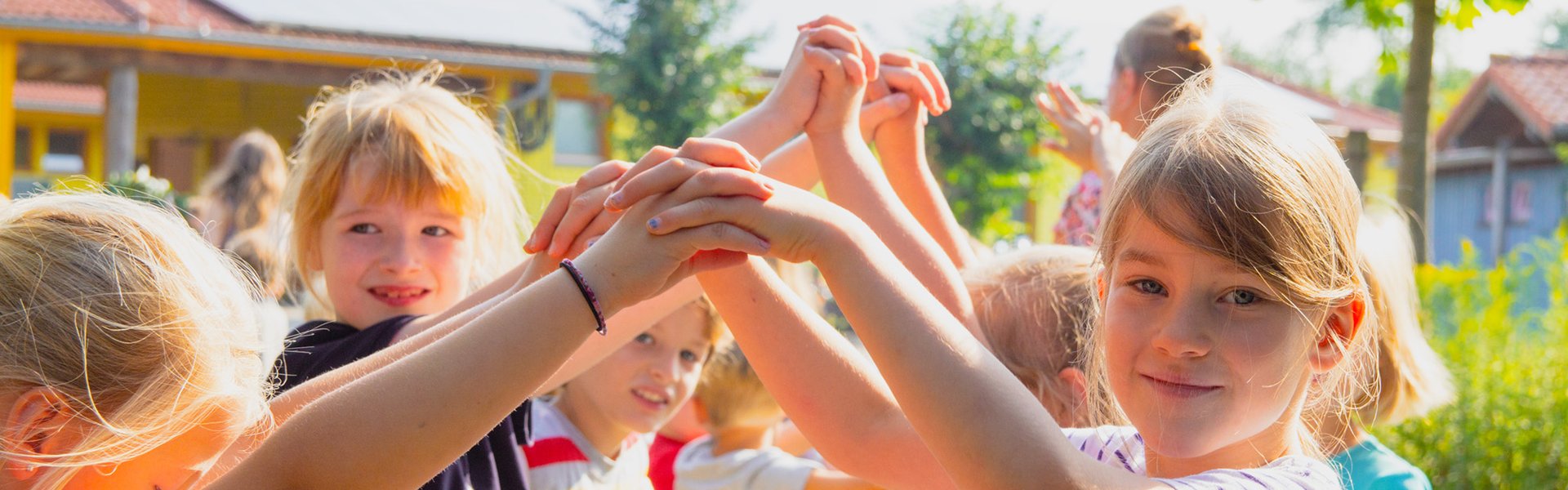 Kinder spielen draußen, lächeln und halten sich an den Händen, während sie im Kreis stehen; im Hintergrund sind Bäume und Gebäude zu sehen – an einem sonnigen Tag.