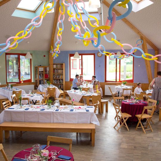 Bunte Papierketten hängen von der Decke eines hellen, festlich geschmückten Raums, in dem Menschen um Tische herum sitzen und stehen, die für eine festliche Veranstaltung oder Feier gedeckt sind. Oberlichter und große Fenster sorgen für Tageslicht.