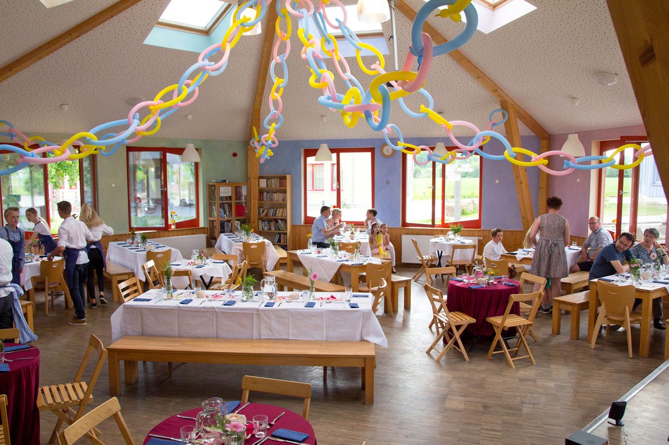 Bunte Papierketten hängen von der Decke eines hellen, festlich geschmückten Raums, in dem Menschen um Tische herum sitzen und stehen, die für eine festliche Veranstaltung oder Feier gedeckt sind. Oberlichter und große Fenster sorgen für Tageslicht.