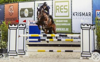 Ein Pferd und sein Reiter springen bei einem Springreiten-Wettbewerb über ein Hindernis, im Hintergrund sind Werbebanner zu sehen.
