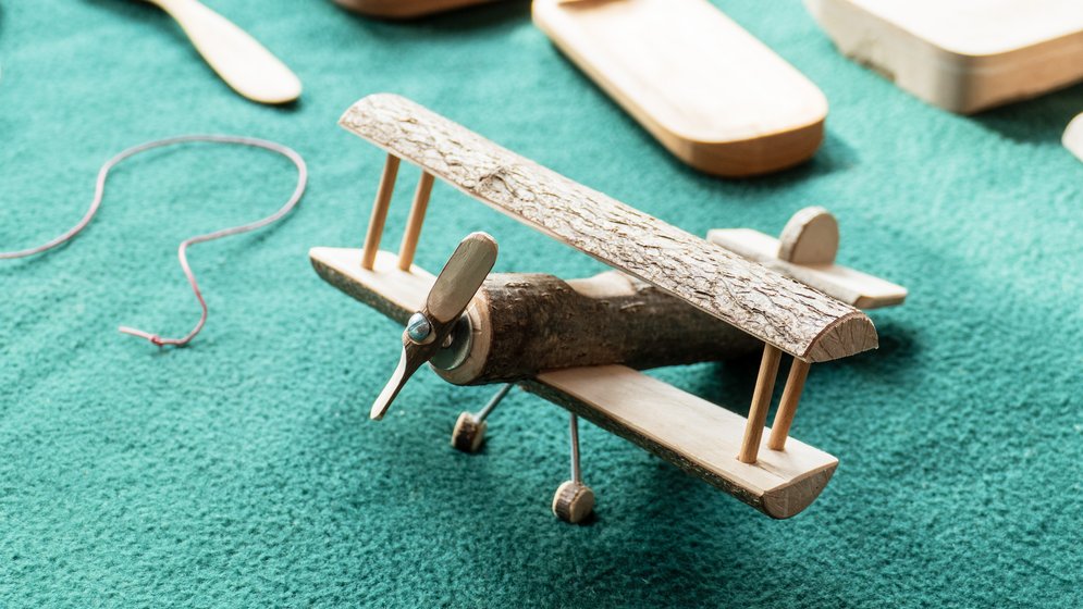 Ein handgefertigtes Spielzeug-Doppeldecker aus Holz, hergestellt aus einem Ast und Holzstücken, steht auf einer strukturierten grünen Fläche, im Hintergrund sind Holzschalen und eine Schnur zu sehen.