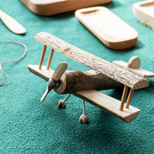 Ein handgefertigtes Spielzeug-Doppeldecker aus Holz, hergestellt aus einem Ast und Holzstücken, steht auf einer strukturierten grünen Fläche, im Hintergrund sind Holzschalen und eine Schnur zu sehen.