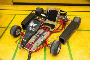 Ein kleines rot-schwarzes Go-Kart mit einem einzigen Sitz steht auf dem gelben Boden einer Sporthalle, der mit grünen Linien markiert ist. Das Go-Kart verfügt über ein Lenkrad und Seitenschutzbügel.