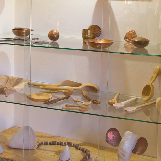 Auf den Glasregalen eines Schranks sind Schalen, Löffel und Küchenutensilien aus Holz und Metall ausgestellt. Auf dem Holzsockel darunter liegen glatte Steine und ein weißes, geformtes Objekt. Eine warme Beleuchtung bringt die handgefertigten Gegenstände besonders gut zur Geltung.
