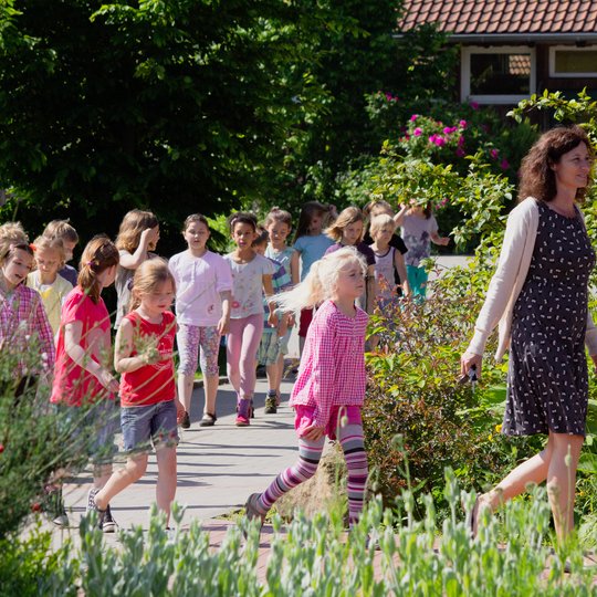 An einem sonnigen Tag läuft eine Gruppe kleiner Kinder draußen in einer Reihe hinter einer Frau in einem schwarzen Kleid mit weißen Mustern her. Entlang des Weges stehen grüne Pflanzen und Blumen.