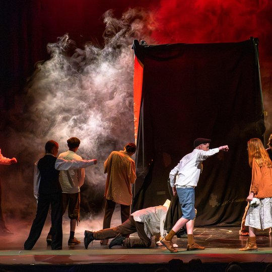 Schauspieler in historischen Kostümen spielen auf der Bühne eine dramatische Szene mit Nebeleffekten, sattrotem Licht und einem dunklen Hintergrund, während ein Schauspieler scheinbar die Hand nach einem anderen ausstreckt oder auf ihn zeigt.