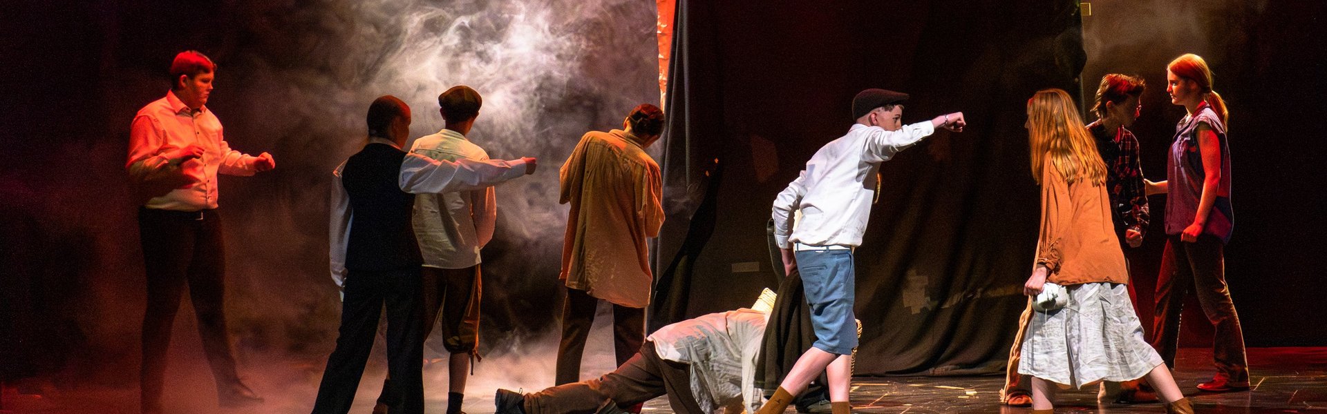 Schauspieler in historischen Kostümen spielen auf der Bühne eine dramatische Szene mit Nebeleffekten, sattrotem Licht und einem dunklen Hintergrund, während ein Schauspieler scheinbar die Hand nach einem anderen ausstreckt oder auf ihn zeigt.