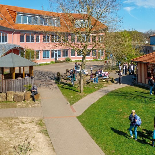 Ein Schulhof, auf dem Schüler in kleinen Gruppen stehen und sich unterhalten. Unter einem strahlend blauen Himmel sind Bäume, Rasenflächen und zwei Gebäude mit roten Dächern zu sehen.