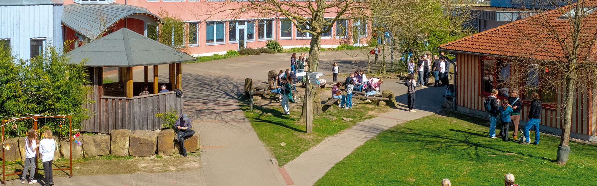 Ein Schulhof, auf dem Schüler in kleinen Gruppen stehen und sich unterhalten. Unter einem strahlend blauen Himmel sind Bäume, Rasenflächen und zwei Gebäude mit roten Dächern zu sehen.