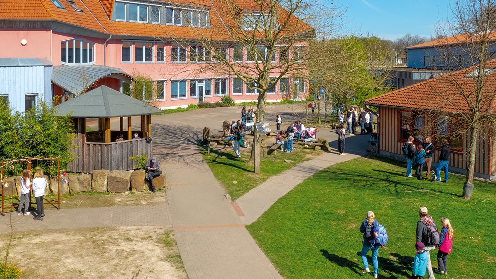 Ein Schulhof, auf dem Schüler in kleinen Gruppen stehen und sich unterhalten. Unter einem strahlend blauen Himmel sind Bäume, Rasenflächen und zwei Gebäude mit roten Dächern zu sehen.