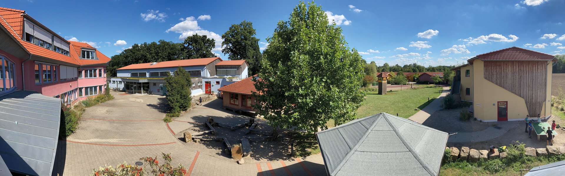 Panoramablick auf einen Schulhof mit Gebäuden mit roten Dächern, grünen Bäumen, einem Spielplatz und spielenden Kindern unter einem sonnigen, blauen Himmel mit vereinzelten Wolken.