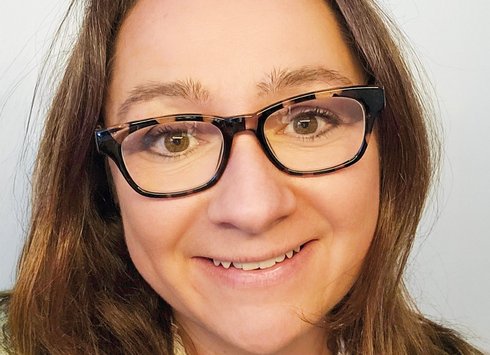 Eine Frau mit braunen Haaren und Brille lächelt in die Kamera; sie trägt ein weißes Oberteil und einen bunten Pullover. Der Hintergrund ist einfarbig und hell.
