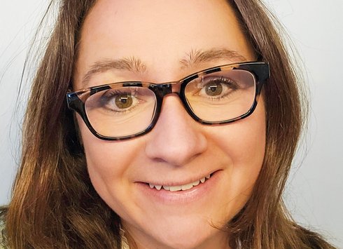 Eine Frau mit braunen Haaren und Brille lächelt in die Kamera; sie trägt ein weißes Oberteil und einen bunten Pullover. Der Hintergrund ist einfarbig und hell.