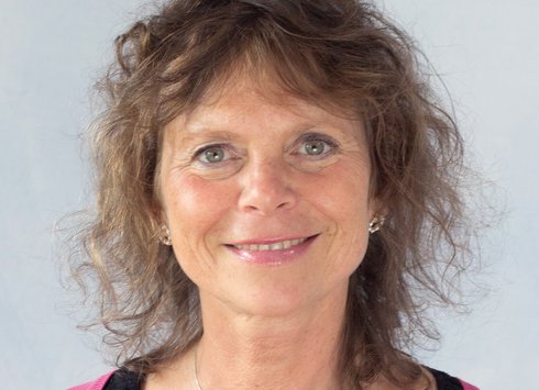 Eine Frau mit lockigem braunem Haar und grünen Augen lächelt in die Kamera. Sie trägt eine rosa Strickjacke, ein schwarzes Oberteil und eine silberne Halskette. Der Hintergrund ist schlicht und hell.
