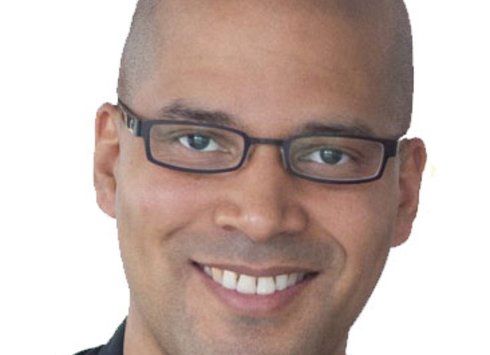 Vor einem schlichten weißen Hintergrund ist eine lächelnde Person mit rasiertem Kopf zu sehen, die eine rechteckige Brille und ein dunkles Hemd trägt.