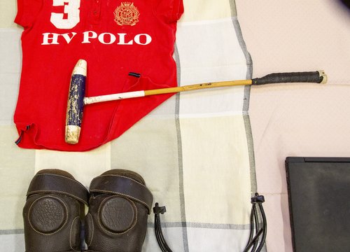 Rotes Polohemd mit dem Schriftzug „3 HV POLO“, einem Poloschläger, braunen Knieschonern, einem schwarzen Kragen und einem schwarzen, rechteckigen Element, die ordentlich auf einem karierten Stoff angeordnet sind.