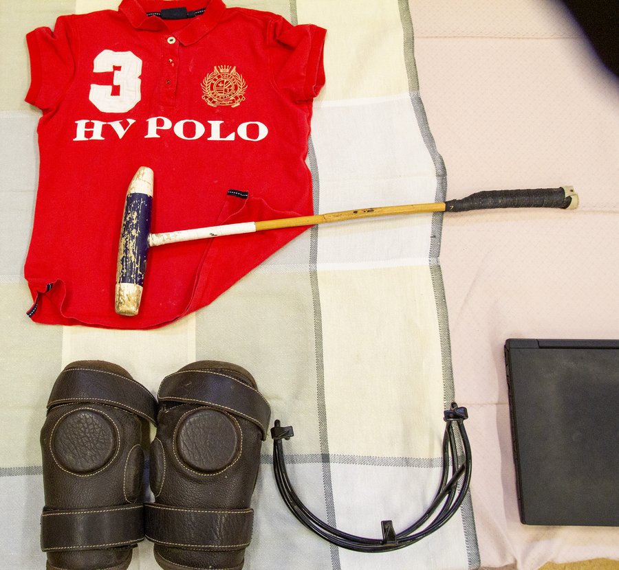 Rotes Polohemd mit dem Schriftzug „3 HV POLO“, einem Poloschläger, braunen Knieschonern, einem schwarzen Kragen und einem schwarzen, rechteckigen Element, die ordentlich auf einem karierten Stoff angeordnet sind.