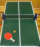 Eine kleine grüne Tischtennisanlage mit einem Netz, einem roten Schläger und einem gelben Ball, der auf dem Tisch liegt. Die Szene spielt in einem Innenraum, und der Tisch hat weiße Begrenzungslinien.