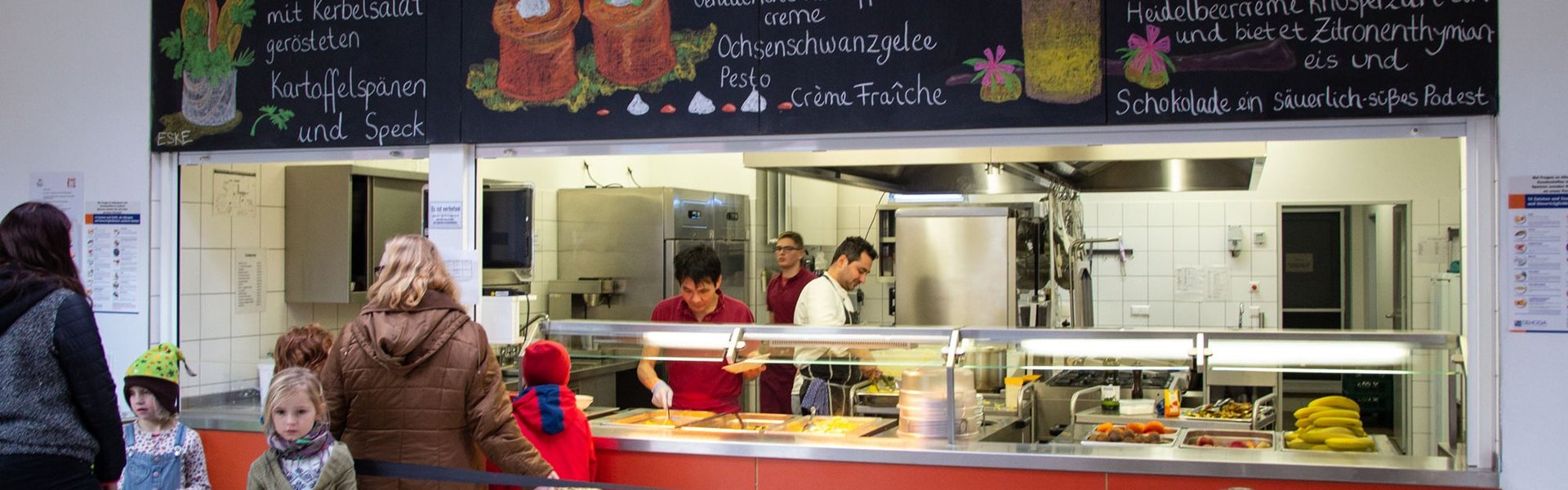Kinder und Erwachsene warten an der Theke einer Cafeteria, wo das Personal das Essen ausschenkt. Über ihnen hängen bunte Dekorationen, und die Gerichte auf der Speisekarte sind auf deutschen Kreidetafeln aufgeschrieben. In der Küche und im Ausgabebereich herrscht reges Treiben, und alles ist sauber.