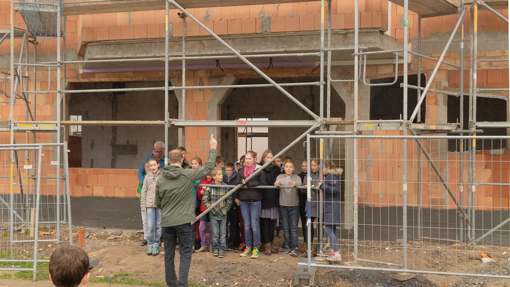 Eine Gruppe von Menschen, darunter auch Kinder, steht vor einem noch nicht fertiggestellten Backsteingebäude mit Gerüsten, während andere oben auf dem Bauwerk stehen. Im Vordergrund beobachten mehrere Kinder das Geschehen von hinten.