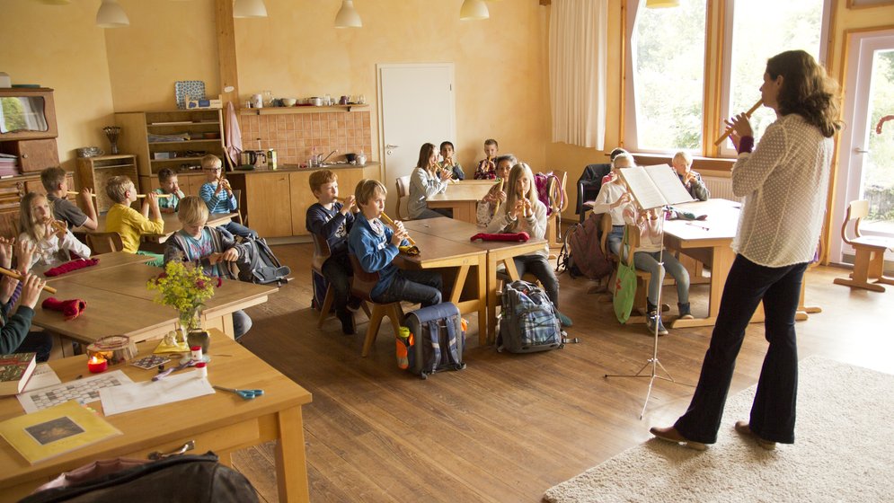 Ein Lehrer spielt auf einer Blockflöte, während die Schüler an ihren Schreibtischen sitzen und ihm in einem hellen Klassenzimmer mit Holzböden und großen Fenstern zusehen und zuhören.