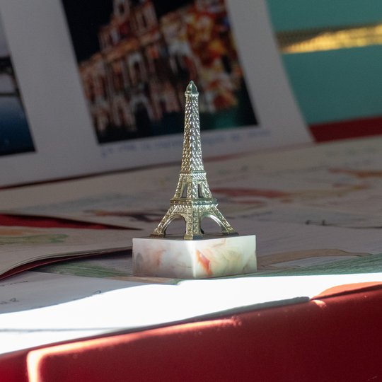 Eine kleine Eiffelturm-Figur steht auf einem Marmorsockel, umgeben von Papieren und Fotos auf einer roten Fläche, während Sonnenlicht die Szene in den Glanz taucht.